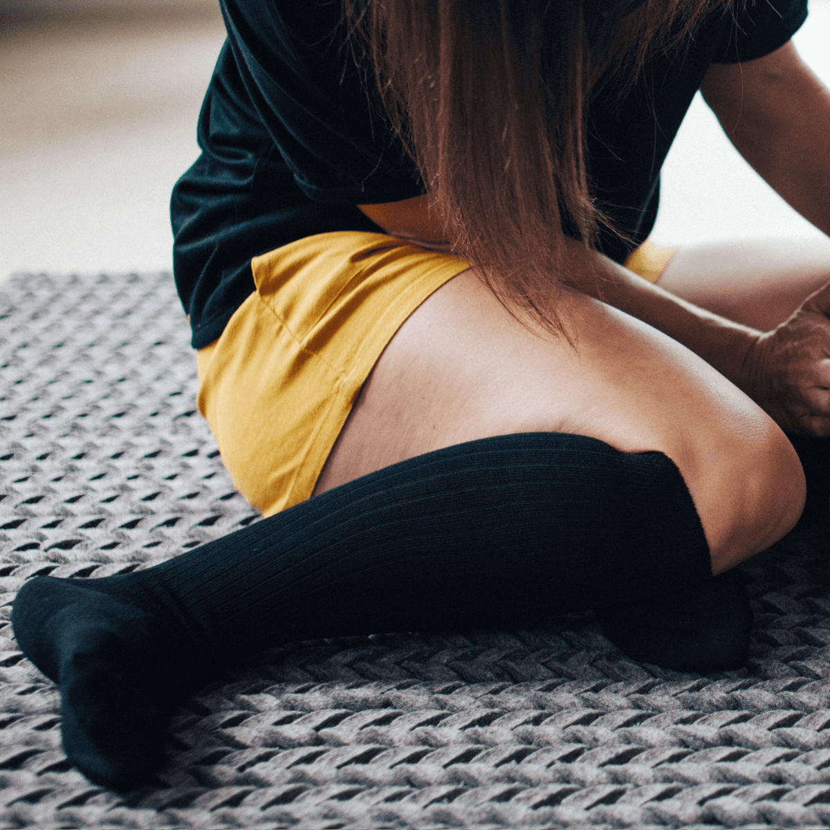Merino Wool Knee High Socks in Black worn by a woman sitting on a textured rug, showcasing a stylish, essential look.